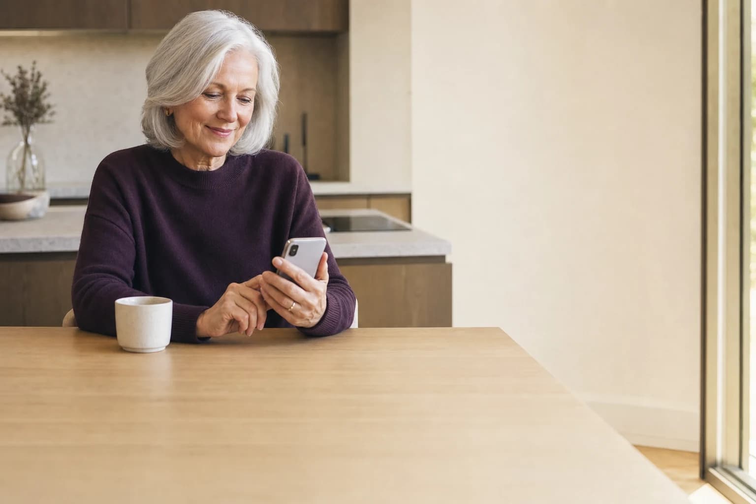 A mother in her seventies at her sunlit kitchen table, reading a note from her family on her phone